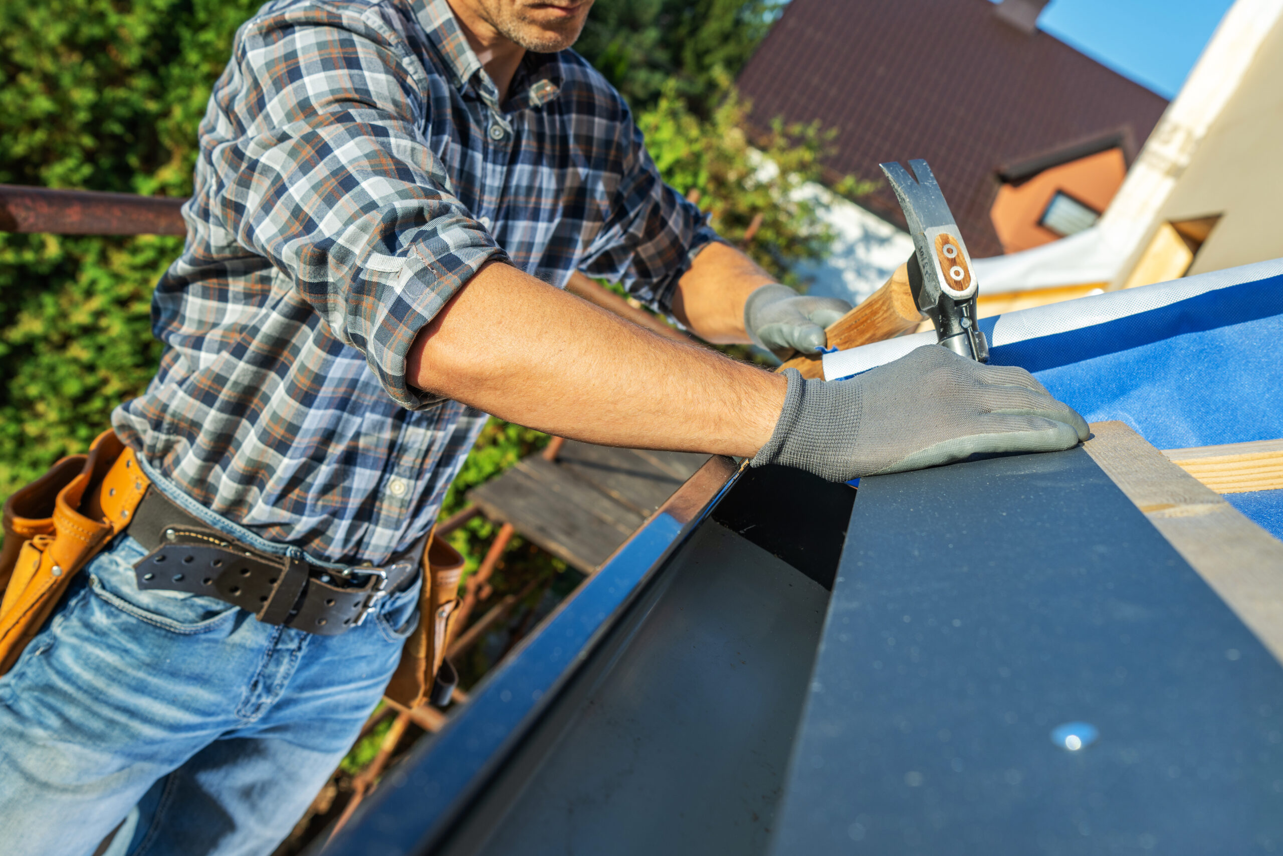 A carpenter diligently installs metal roofing on a residential building. The worker wears protective gloves and a plaid shirt, focusing on precision while using a hand tool.